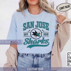 May include: Light blue t-shirt featuring the text "SAN JOSE Sharks" with a logo of a shark and hockey sticks. The shirt includes the text "EST. 2019" and the shop's name. The words "LIGHT BLUE" and "CUSTOM YEAR" are also visible.