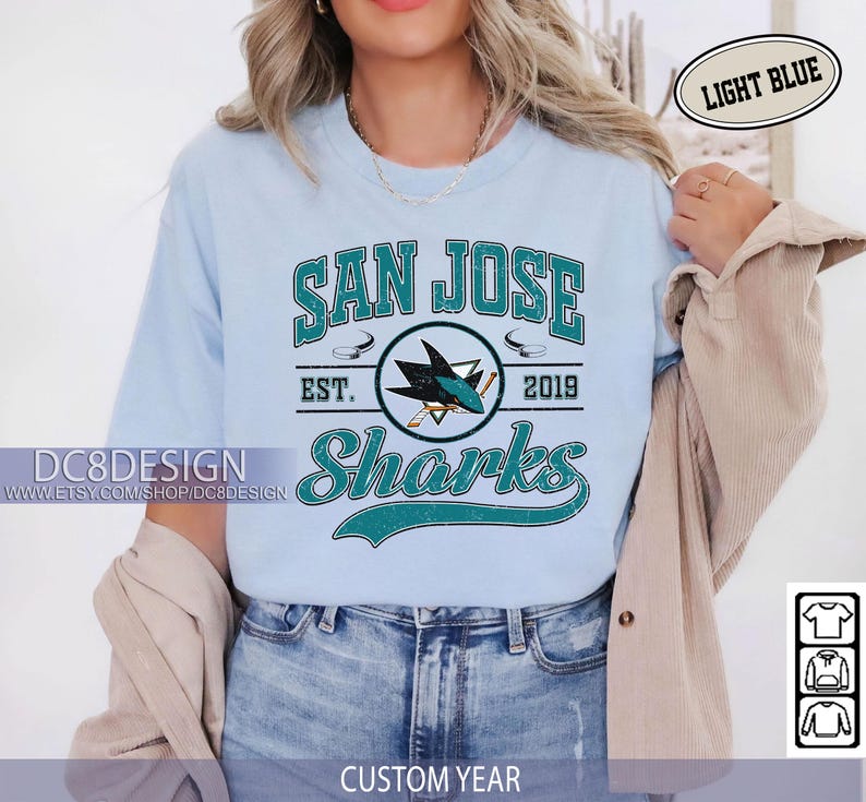 May include: Light blue t-shirt featuring the text "SAN JOSE Sharks" with a logo of a shark and hockey sticks. The shirt includes the text "EST. 2019" and the shop's name. The words "LIGHT BLUE" and "CUSTOM YEAR" are also visible.