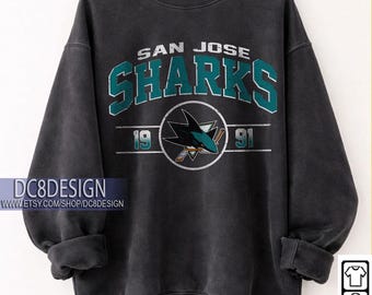 May include: Black San Jose Sharks sweatshirt with teal and white lettering and logo. The sweatshirt features the text "SAN JOSE SHARKS" and the years "19" and "91". The sweatshirt is hanging on a wooden hanger. The text "BLACK" is in a black oval.