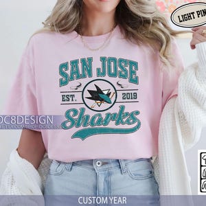 May include: Light pink t-shirt featuring the San Jose Sharks logo in teal and white. The design includes the text "SAN JOSE Sharks" and "EST. 2019". The shirt also has the text "DC8DESIGN" and "CUSTOM YEAR".