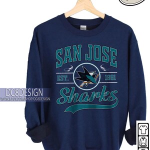 May include: Navy blue sweatshirt featuring the text "SAN JOSE Sharks" in teal and white. The design includes a shark logo and the year "1991". The sweatshirt is hanging on a wooden hanger.