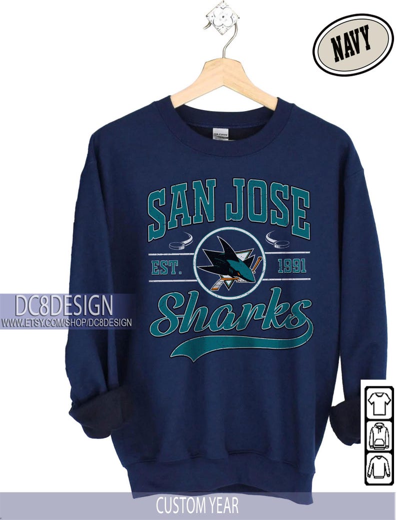 May include: Navy blue sweatshirt featuring the text "SAN JOSE Sharks" in teal and white. The design includes a shark logo and the year "1991". The sweatshirt is hanging on a wooden hanger.