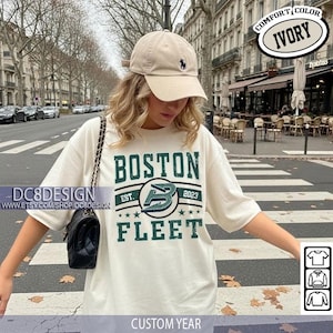 Può includere: T-shirt color crema con testo "BOSTON FLEET" in verde scuro. La maglietta presenta un logo con la scritta "EST. 2023" e stelle. Viene indossato un berretto da baseball beige con logo nero. L'immagine include anche una borsa nera.