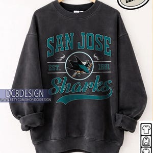 Könnte beinhalten: Schwarzes San Jose Sharks Sweatshirt mit teal- und weißfarbenem Schriftzug und Logo. Das Design beinhaltet "SAN JOSE" über "Sharks" und das Jahr "1991". Das Sweatshirt wird auf einem Kleiderbügel präsentiert.