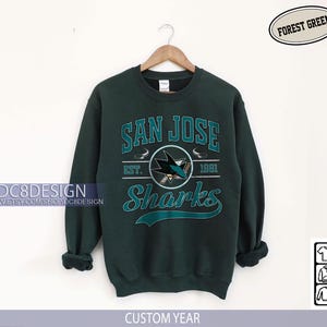 May include: Forest green San Jose Sharks sweatshirt. The sweatshirt features the team name and logo in teal and white, with the text "San Jose Sharks" and "Est. 1991". The sweatshirt is hanging on a wooden hanger.