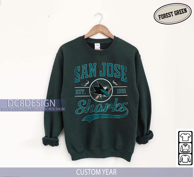 May include: Forest green San Jose Sharks sweatshirt. The sweatshirt features the team name and logo in teal and white, with the text "San Jose Sharks" and "Est. 1991". The sweatshirt is hanging on a wooden hanger.