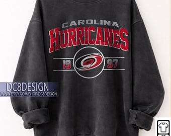 May include: Black Carolina Hurricanes sweatshirt with red and white team logo and text. The design includes the team name, logo, and the year 1997. The sweatshirt is displayed on a wooden hanger.