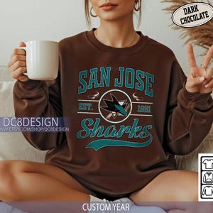 May include: Dark chocolate brown San Jose Sharks sweatshirt with teal lettering and logo. The sweatshirt features the words "San Jose Sharks" and "Est. 1991". A white mug is held in the left hand.