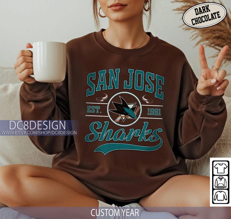 May include: Dark chocolate brown San Jose Sharks sweatshirt with teal lettering and logo. The sweatshirt features the words "San Jose Sharks" and "Est. 1991". A white mug is held in the left hand.