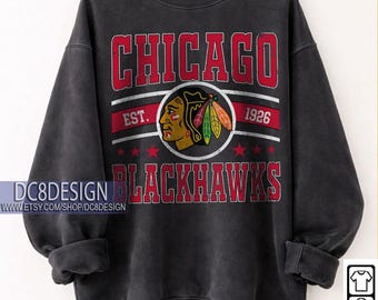 May include: A black crewneck sweatshirt featuring a distressed graphic with the text CHICAGO BLACKHAWKS EST. 1926. The design includes a central circular emblem of a Native American profile with feathers and red stars. The text DC8DESIGN is visible.