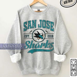May include: Ash gray sweatshirt with "SAN JOSE Sharks" text and logo in teal and white. The design includes the text "EST. 2019" and a hockey puck graphic. The sleeves are cuffed with white fabric. The text "CUSTOM YEAR" is at the bottom.