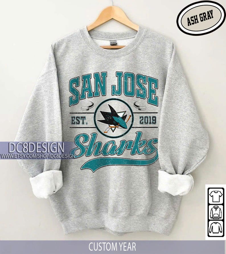 May include: Ash gray sweatshirt with "SAN JOSE Sharks" text and logo in teal and white. The design includes the text "EST. 2019" and a hockey puck graphic. The sleeves are cuffed with white fabric. The text "CUSTOM YEAR" is at the bottom.