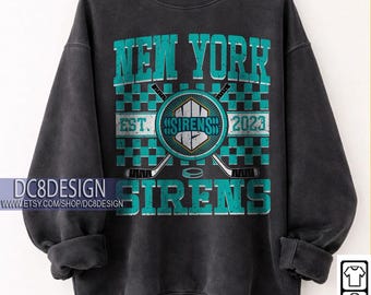 May include: Black crewneck sweatshirt with teal and white text that reads "NEW YORK SIRENS" and "EST. 2023". The design includes crossed hockey sticks, a hockey puck, and a shield emblem. The sweatshirt is hanging on a wooden hanger.