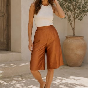 May include: A sleeveless cream-colored ribbed tank top paired with high-waisted, knee-length rust-colored culottes featuring a smooth finish and a clean, tailored silhouette, worn with simple brown strappy sandals.