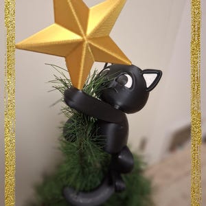 Könnte beinhalten: Ein skurriler Weihnachtsbaumschmuck mit einer schwarzen Katzenfigur, die einen goldenen Stern hält. Die Katze sitzt auf einem kleinen, grünen Kunstbaum. Der Stern ist goldfarben, und das Design ist festlich und verspielt.