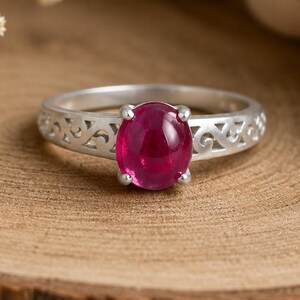 May include: A silver ring with a deep red, oval gemstone. The ring band has a decorative, openwork pattern. The ring is presented on a wooden surface, with a soft, neutral-coloured fabric in the background. A sprig of small, white flowers is also visible.