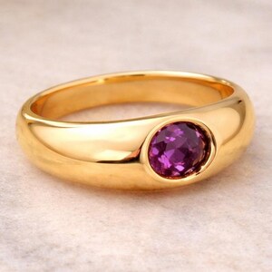 Könnte beinhalten: Ein goldener Ring mit poliertem Finish, der einen leuchtenden lila Edelstein aufweist. Das Design des Rings ist schlicht, aber elegant, mit einem glatten Band und einer Krappenfassung für den Edelstein. Der Ring wird auf einer hellen Oberfläche präsentiert.