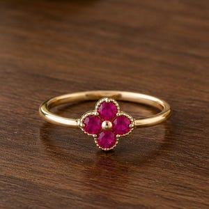 Può includere: Un anello d'oro con un design a quadrifoglio. Il quadrifoglio è composto da quattro pietre preziose rotonde di colore rosso vivo. L'anello è appoggiato su una superficie di legno scuro, con uno sfondo sfocato di verde e una ciotola di ceramica.