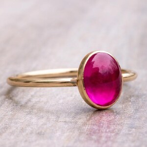 Op de afbeelding: Een gouden ring met een grote, ovaalvormige, helder roze edelsteen. De ring heeft een slanke band. De edelsteen is gezet in een gouden bezel. De ring wordt getoond op een lichtgekleurd oppervlak. Sieraad.