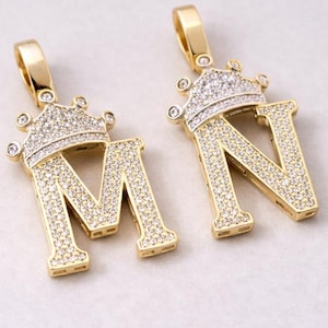 Puede incluir: Dos colgantes dorados con letras "M" y "N" coronadas. Las letras y las coronas están incrustadas con pequeñas piedras preciosas transparentes. Los colgantes tienen una argolla dorada para una cadena.