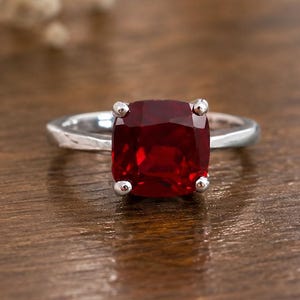 May include: A silver ring with a large, square-cut, deep red gemstone. The ring's band is smooth, and the gemstone is held in place by four small prongs. The ring is set against a warm, wooden surface.