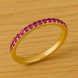 Peut inclure: Une bague dorée ornée d'une rangée de petites pierres précieuses roses vives. La bague est présentée sur une surface en bois brun clair, mettant en valeur le design et la couleur du bijou.