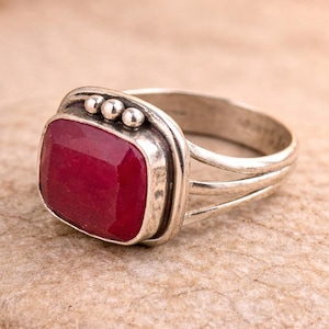 Könnte beinhalten: Ein silberner Ring mit einem großen, rechteckigen, tiefroten Edelstein. Der Ring hat ein schlichtes Band mit geteiltem Design und eine dekorative Fassung um den Stein, mit drei kleinen Silberkügelchen. Der Ring wird auf einem hellbraunen Stein präsentiert.
