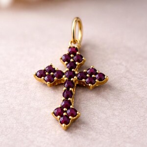 May include: A gold-colored pendant in the shape of a cross, adorned with numerous small, deep red gemstones. The cross has a loop at the top for a chain. The gemstones are set closely together, creating a rich, textured appearance.