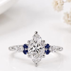 May include: A silver-coloured ring featuring a large, central marquise-cut clear stone. The band is accented with two small, round blue stones on each side of the centre, followed by a smaller marquise-cut clear stone on each outer edge of the setting.