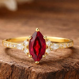 Puede incluir: Un anillo de oro con una piedra preciosa roja oscura con corte marquesa como pieza central. La banda está adornada con pequeñas piedras preciosas claras y el engaste presenta piedras blancas adicionales. El anillo está sobre una superficie de madera rústica.
