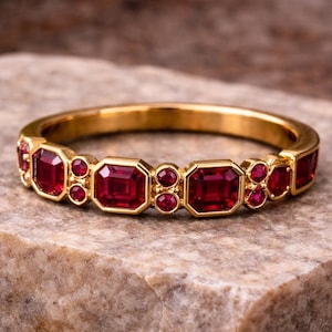 May include: A gold ring with a series of vibrant red gemstones. The ring features a repeating pattern of rectangular and round stones, set in a polished gold band. The ring is displayed on a textured stone surface.