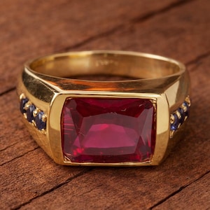 Può includere: Un anello d'oro con una grande pietra preziosa rettangolare rosso rubino. L'anello ha una fascia d'oro con piccole pietre preziose blu scuro su ogni lato della pietra principale. L'anello è appoggiato su una superficie di legno.