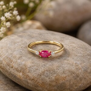 May include: A gold ring featuring a vivid pink oval gemstone, with two small, clear, round gemstones either side. The ring is displayed on a grey stone, with white flowers in the background.