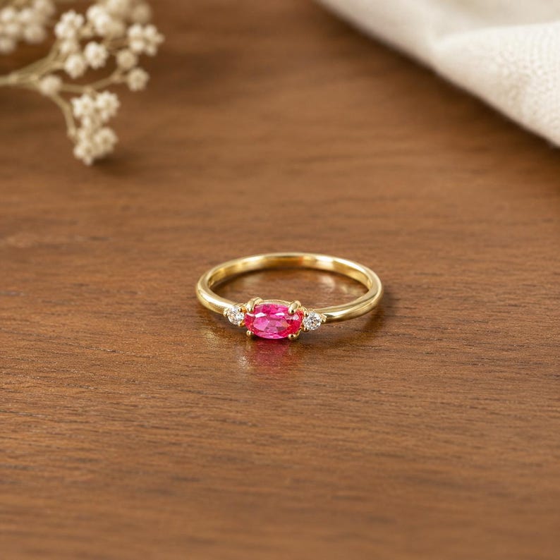 May include: A gold ring featuring a central oval-cut pink gemstone, with two smaller, clear gemstones either side. The ring is displayed on a wooden surface. The background is soft and blurred.