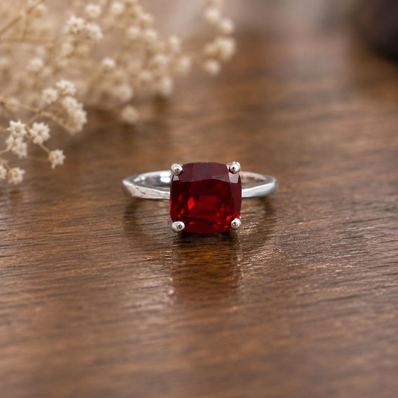May include: A silver ring with a large, square-cut, deep red gemstone. The ring's band is smooth, and the gemstone is held in place by four small prongs. The ring is set against a warm, wooden surface.