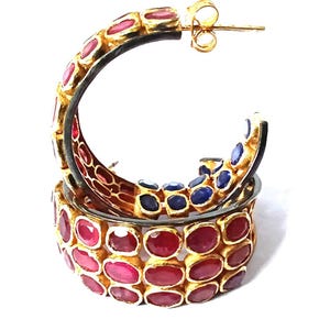 Pode incluir: Um par de brincos de argola dourados. Os brincos são incrustados com pedras preciosas vermelhas e azuis em forma oval. As pedras preciosas são fixadas em uma moldura de metal dourado. Os brincos têm um fecho de pino.