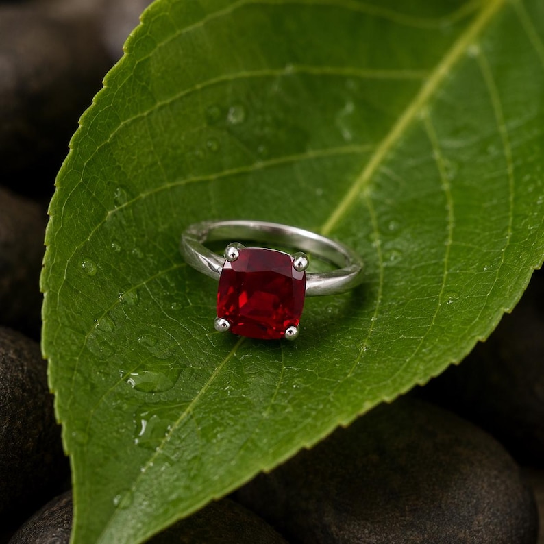 May include: A silver ring with a large, square, deep red gemstone. The ring is set on a green leaf with water droplets, creating a natural and elegant display. The gemstone is approximately 0.5 inches in size.