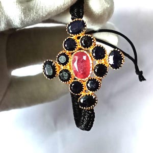 May include: A cross-shaped pendant featuring a central oval ruby surrounded by black and blue gemstones. The pendant is set in gold and attached to a black cord. The design is intricate and elegant.