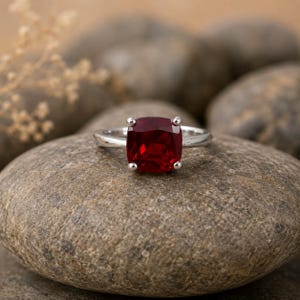 May include: A silver ring featuring a large, square-cut, deep red gemstone. The gemstone is set with four prongs, and the ring is displayed on a gray stone, with other stones in the background. The ring is a piece of jewelry.