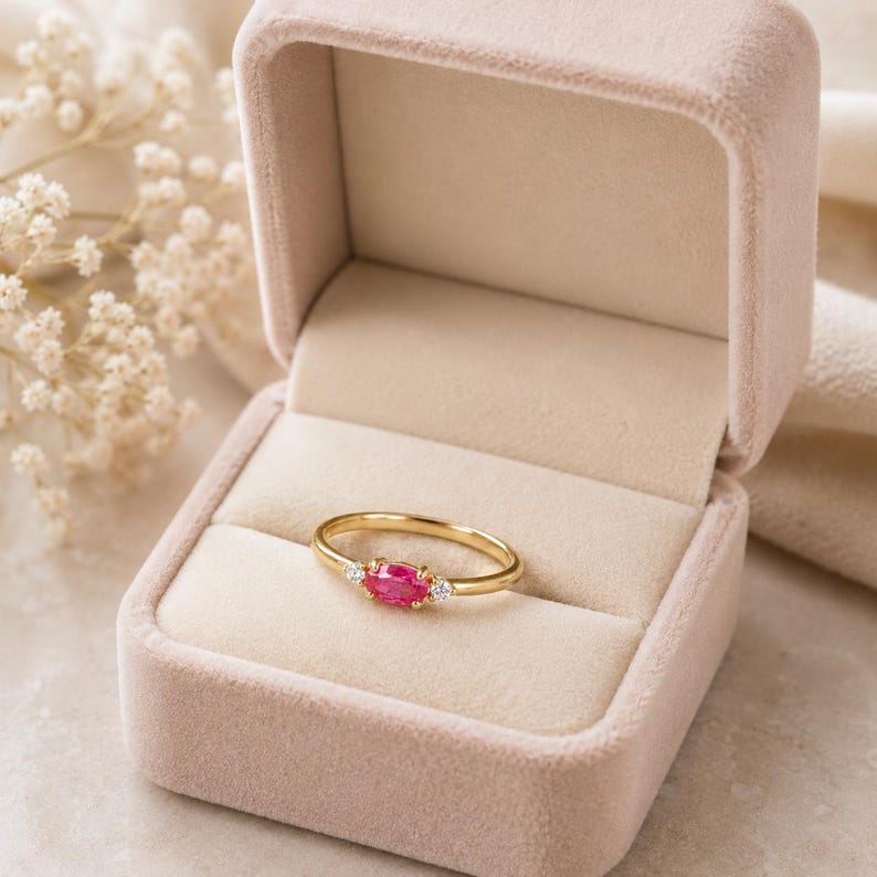 May include: A gold ring featuring a ruby-coloured oval gemstone and two small white gemstones. The ring is presented in an open pink velvet ring box. The box displays the ring against a soft, neutral background.