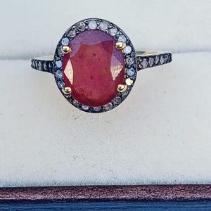 Pode incluir: Um close-up de um anel com uma grande pedra preciosa vermelha em forma oval. A pedra preciosa é rodeada por um halo de pequenas pedras transparentes. A banda é preta com pequenas pedras transparentes. O anel tem uma cravação em ouro.
