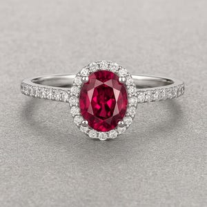Puede incluir: Un anillo de plata con una gran piedra preciosa ovalada de color rojo intenso en el centro, rodeada por un halo de piedras claras más pequeñas. La banda del anillo también está engastada con pequeñas piedras claras, creando un efecto brillante. Fondo gris.