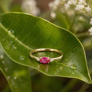 May include: A gold ring featuring a central oval pink gemstone, flanked by two smaller white stones. The ring rests on a green leaf with water droplets, with white flowers in the background.