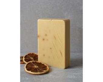 May include: A rectangular bar of pale yellow soap with small brown spots and streaks. Two dried orange slices are in front of the soap. The soap and oranges are on a grey surface.