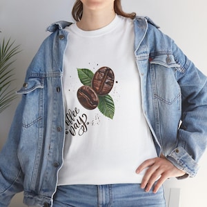 May include: White t-shirt with a coffee-themed design. The graphic shows two roasted coffee beans, green leaves, and the words "Coffee Day" in a script font. The shirt is worn with a denim jacket.