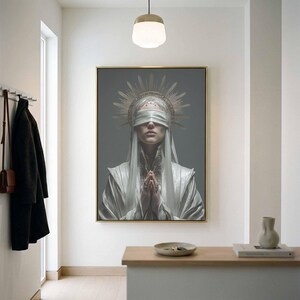 May include: A rectangular wall art print in a gold frame featuring a portrait of a figure wearing a white blindfold, a metallic sunburst headpiece, and a white robe with hands pressed together in a prayer position against a solid gray background.