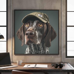 May include: A square wall art print featuring a portrait of a brown and white speckled dog wearing an olive green baseball cap. The dog has amber eyes and long, dark brown ears. The background is a solid, neutral gray color within a black frame.