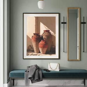 May include: A framed wall art print depicting three terracotta-colored earthenware vessels in a sunlit corner, displayed above a teal velvet bench with a grey coat, a white handbag, and black gloves resting on the seat next to a tall mirror.