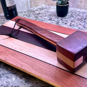 May include: A rectangular wooden cutting board featuring alternating stripes of light, medium, and dark brown wood grain. Resting on top is a wooden mallet with a matching multi-toned wood handle and head, equipped with a thin black leather hanging cord.