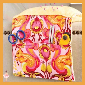 May include: A fabric armchair caddy with a pink, orange, and yellow squirrel and floral pattern. It features a light yellow padded top with pins, two front pockets holding blue-handled scissors, a metal ruler, and a yellow rotary cutter.